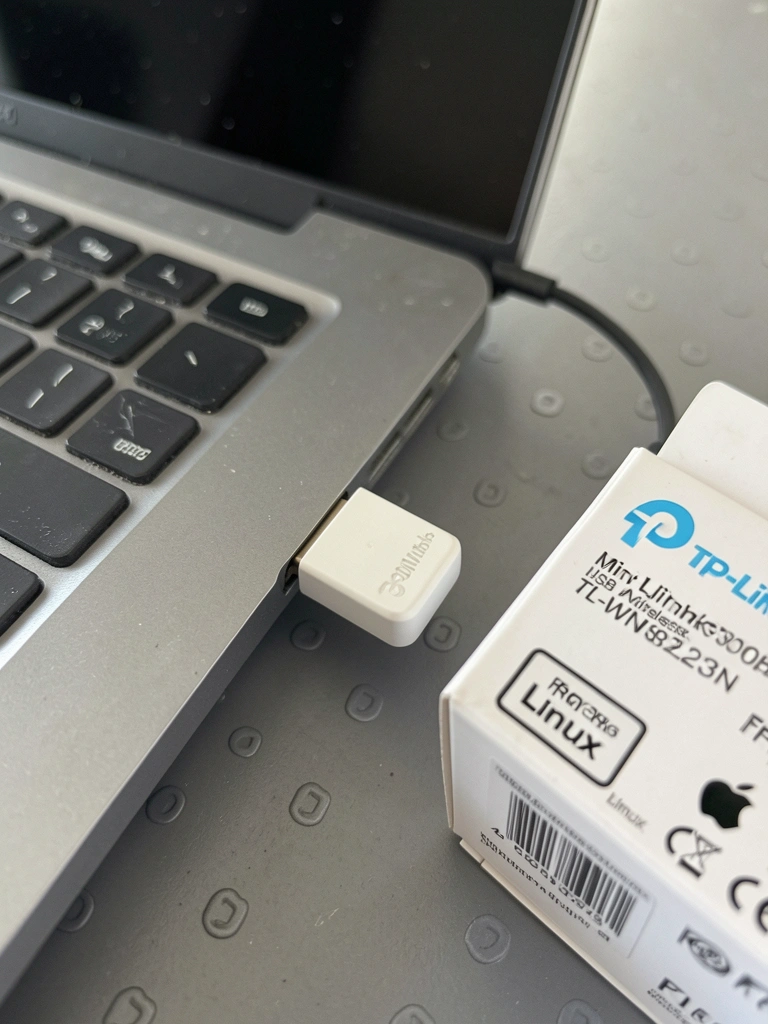 Dongle Wi-Fi USB prateado conectado a um laptop prateado sobre uma mesa de madeira.