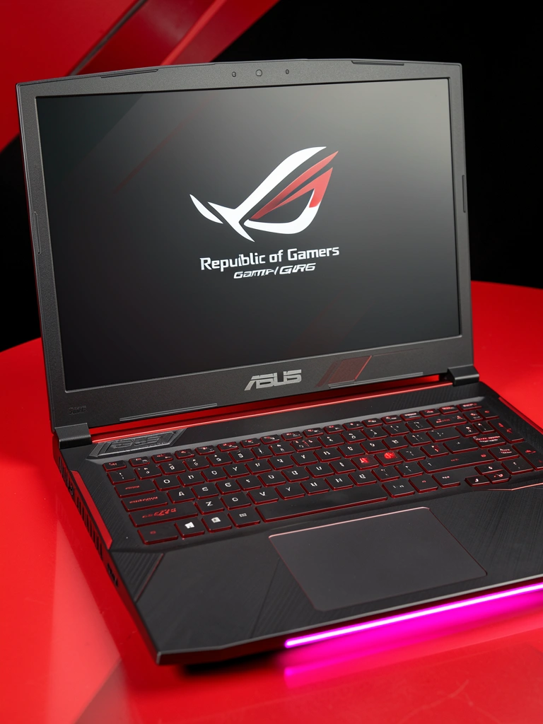 Notebook ASUS ROG Strix G513 preto com detalhes vermelhos aberto sobre superfície preta e vermelha, faixa de LED rosa sob a base, tela escura com logotipo Republic of Gamers em branco.