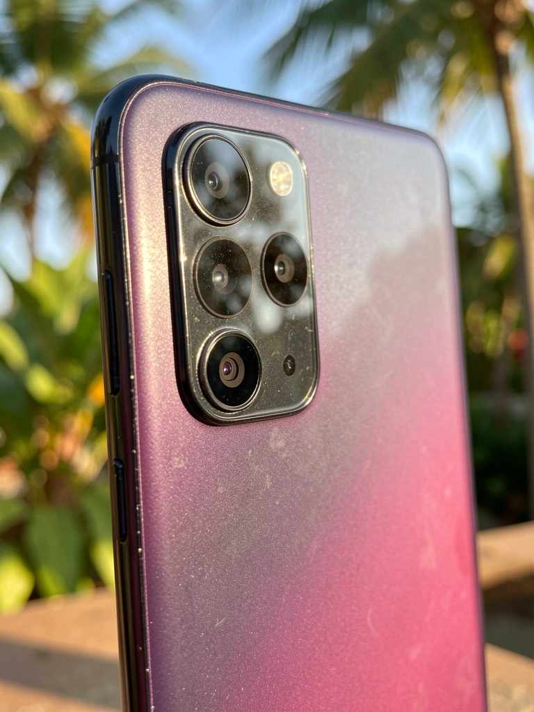 Close-up vertical de smartphone Samsung Galaxy S23 preto com gradiente roxo e rosa, câmera tripla e flash, luz natural quente e fundo desfocado com folhagens.